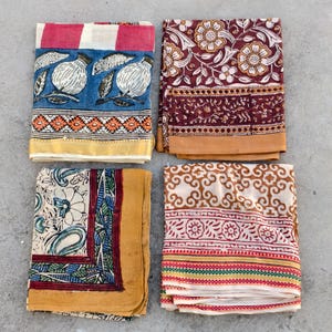 May include: Four folded textiles with diverse patterns. One features blue with white bird illustrations, another has a floral design on a burgundy background, a third has a floral and paisley design, and the last has a repeating pattern with red and yellow stripes.