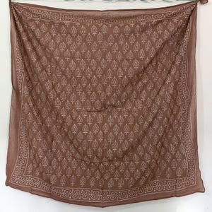 Puede incluir: Bufanda marrón con un patrón repetitivo de gotas blancas. La bufanda tiene un borde decorativo con un diseño floral blanco. La bufanda está colgada contra una pared blanca.