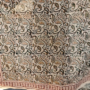 May include: A beige textile with a repeating paisley pattern in shades of green, brown, and black. The border features a repeating pattern of small circles in a deep red colour. The textile is likely a scarf or shawl.