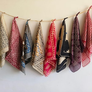 May include: A display of patterned fabric scarves in a range of colours, including green, red, blue, and black, suspended from a string against a white wall. Each scarf showcases a unique geometric or floral design, creating a vibrant textile display.