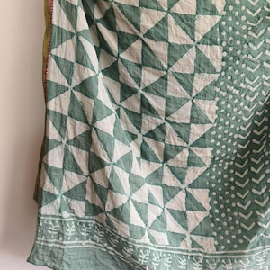 Op de afbeelding: Een gedrapeerd textiel met een geometrisch patroon van witte driehoeken en teal-groene ruiten. De stof heeft een rand met een herhalend bladmotief. De rand is afgewerkt met contrasterende groene en rode stiksels.