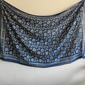 May include: A blue scarf with a white floral pattern. The scarf has a border with a repeating geometric design. The fabric appears to be lightweight and flowing, suitable for warmer weather. The scarf is hanging against a white background.