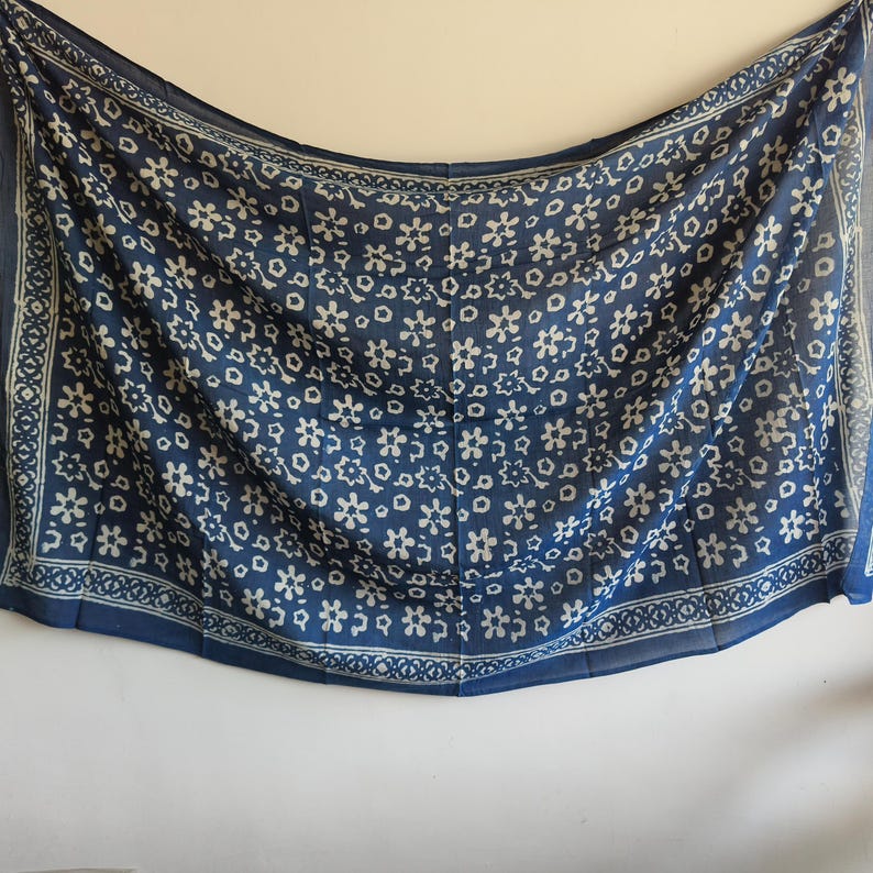 May include: A blue scarf with a white floral pattern. The scarf has a border with a repeating geometric design. The fabric appears to be lightweight and flowing, suitable for warmer weather. The scarf is hanging against a white background.