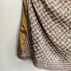 Puede incluir: Bufanda estampada con un dise&ntilde;o floral marr&oacute;n y crema. La bufanda tiene un borde dorado y un borde decorativo. La tela parece ligera y fluida, adecuada para climas c&aacute;lidos.