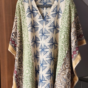 May include: A cream-colored, V-neck tunic with a floral pattern in blue and green. The tunic has wide sleeves and a gold border. The fabric appears to be lightweight, suitable for warm weather.