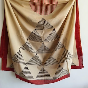 Op de afbeelding: Beige stof met een geometrisch ontwerp. De stof heeft een patroon van zwarte driehoeken en een rode halve cirkel met verticale lijnen. De randen zijn afgewerkt met een effen rode rand. De stof is gedrapeerd en toont zijn textuur en ontwerp.