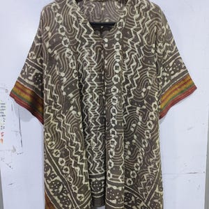 May include: A brown, patterned tunic with a V-neck and short sleeves. The tunic features a repeating white geometric design and a colorful border on the sleeves. The garment is hanging on a black hanger.