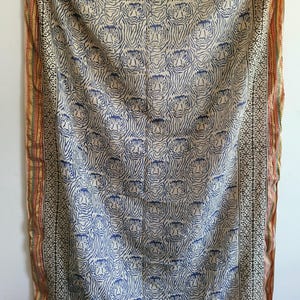 May include: A cream-colored textile with a repeating pattern of blue tiger faces. The fabric has a border with geometric and floral designs in black, red, and gold. The textile is likely a decorative throw or wall hanging.