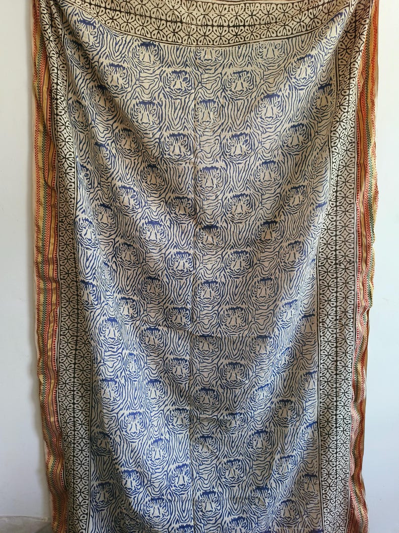 May include: A cream-colored textile with a repeating pattern of blue tiger faces. The fabric has a border with geometric and floral designs in black, red, and gold. The textile is likely a decorative throw or wall hanging.
