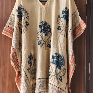 May include: Cream-colored kaftan with a V-neck and short sleeves. The kaftan features large blue floral patterns and decorative borders with geometric and colorful designs. The fabric appears lightweight and flowing, suitable for warm weather.