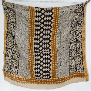 May include: A square scarf with a cream background and a repeating pattern in black, brown, and gold. The scarf features geometric and floral designs, with a gold border. The scarf is hanging from a thin string.