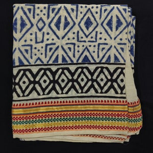 May include: A white cotton fabric with a blue and black geometric pattern. The fabric has a colourful border with red, yellow, green, and gold threads.