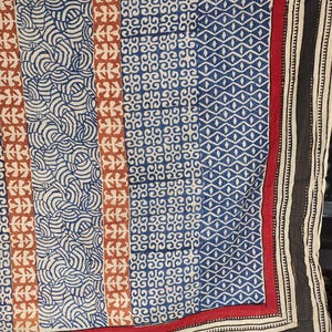May include: A patterned textile with a mix of geometric and organic designs in navy blue, cream, rust, and red. The border is black and white striped. The fabric is likely used for clothing or home decor. The design is intricate and detailed.