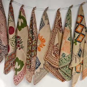 May include: A display of patterned bandanas hanging against a white wall. The bandanas showcase diverse designs, including floral, geometric, and bird motifs, in shades of orange, green, and brown. They are made of a light-coloured fabric.