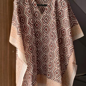 May include: A cream-colored, V-neck caftan with a geometric diamond pattern in shades of burgundy and gray. The edges are trimmed with a cream-colored border featuring a repeating design. The caftan is hanging on a black hanger.