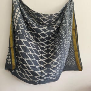 May include: A grey and white patterned scarf with a geometric design. The scarf has a yellow border and a repeating diamond pattern. The fabric appears lightweight and flowing, suitable for various styles. The scarf is draped.