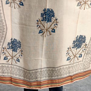 Op de afbeelding: Crèmekleurige stof met een bloemenpatroon in blauw en bruin. Het ontwerp toont grote gestileerde bloemen en gedetailleerde randpatronen in zwart en rood. De stof is waarschijnlijk een textiel, mogelijk voor kleding of woondecoratie.