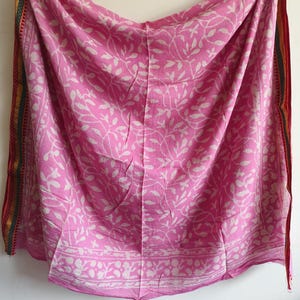 Pode incluir: Um tecido rosa com um padrão de folhas brancas. As bordas são adornadas com bordas coloridas e estampadas. O tecido é drapeado, mostrando o design e a textura. Este tecido pode ser um lenço, um xale ou um tecido decorativo.