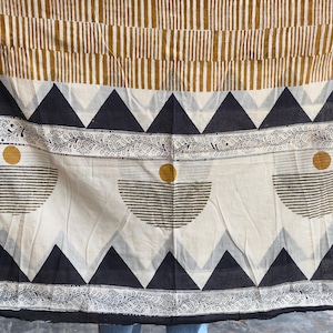 Op de afbeelding: Een textiel met een geometrisch patroon in zwart, wit en goud. Het ontwerp omvat driehoeken, halve cirkels en strepen. De rand is zwart.