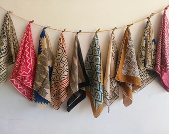 May include: A collection of ten patterned bandanas hanging from a string against a white wall. The bandanas feature various geometric and floral designs in colors like red, blue, black, and tan. Each bandana is secured with a wooden clip.