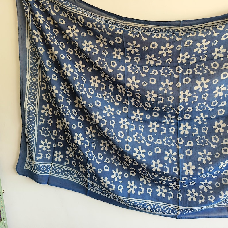 May include: A blue scarf with a white floral pattern and a decorative border. The scarf is made of a lightweight fabric and is draped against a white wall. The floral design is repeated across the scarf's surface.