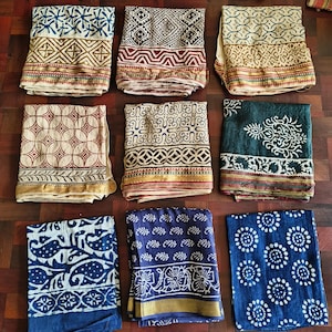 May include: Nine folded textiles with diverse patterns and colours are displayed. The textiles feature geometric and floral designs in shades of blue, red, cream, and green. Each textile has a decorative border.