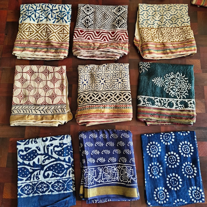 May include: Nine folded textiles with various patterns and colors are displayed. The textiles feature geometric and floral designs in shades of blue, red, cream, and green. Each textile has a decorative border.