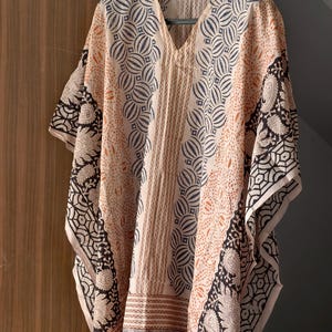 Op de afbeelding: Een beige kaftan met een V-hals en een geometrische en paisley print. De kaftan heeft een mix van patronen in zwart, marineblauw en oranje, met een randontwerp langs de zoom en mouwen. Het kledingstuk hangt aan een zwarte hanger.