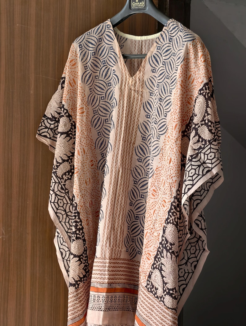 Op de afbeelding: Een beige kaftan met een V-hals en een geometrische en paisley print. De kaftan heeft een mix van patronen in zwart, marineblauw en oranje, met een randontwerp langs de zoom en mouwen. Het kledingstuk hangt aan een zwarte hanger.