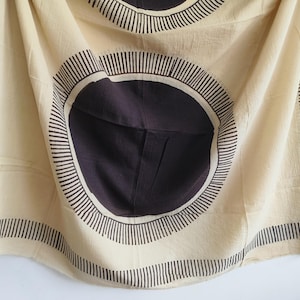 May include: A beige textile with a repeating circular pattern in dark brown and white. The design features large dark brown circles with white outlines and radiating lines, set against a beige background. The edges are bordered with vertical lines.