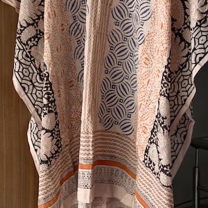 Op de afbeelding: Een cr&egrave;mekleurige kaftan met ingewikkelde patronen in zwart, marineblauw en oranje. Het ontwerp bevat bloemen- en geometrische motieven, met een rand met gedetailleerde patronen. De stof lijkt licht en vloeiend.