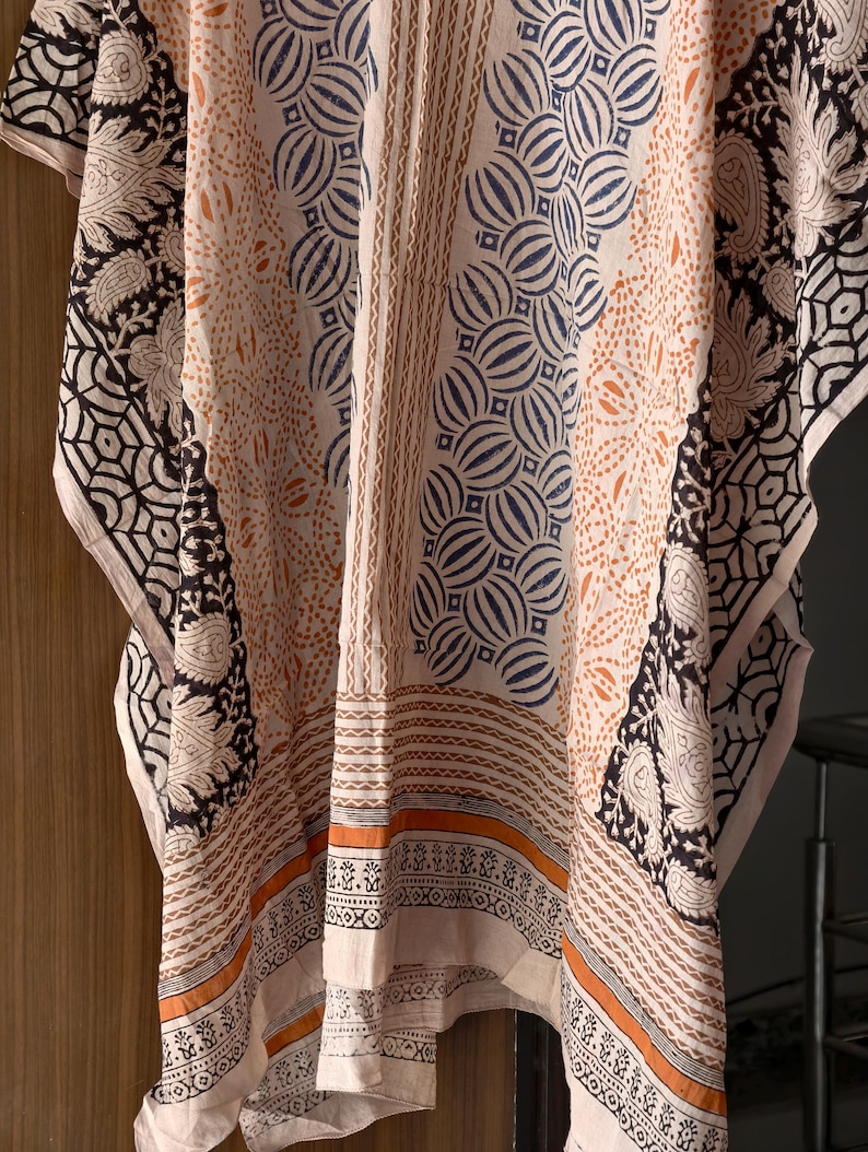 Op de afbeelding: Een cr&egrave;mekleurige kaftan met ingewikkelde patronen in zwart, marineblauw en oranje. Het ontwerp bevat bloemen- en geometrische motieven, met een rand met gedetailleerde patronen. De stof lijkt licht en vloeiend.