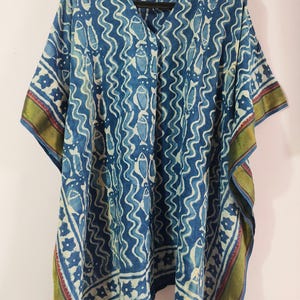 May include: A blue and white patterned kaftan with a V-neck. The fabric features a repeating fish and wave design. The edges are trimmed with a gold, red, and green border. The garment is hanging on a black hanger.