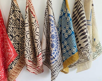 May include: A collection of patterned fabric scarves in various colors, including red, beige, and blue. The scarves feature geometric and floral designs in black and brown, with some having decorative borders. They are hanging against a white background.