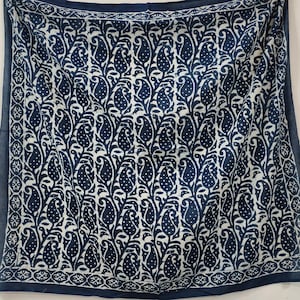 May include: An indigo blue and white scarf with a repeating paisley design. The pattern features stylized teardrop shapes with dotted details, set against a white background. The border has a decorative geometric pattern.