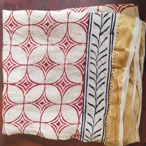 May include: Folded cream-colored fabric with a repeating pattern of red diamonds within circles. A black and white leaf design borders the fabric, with a gold and cream-colored woven edge. The fabric is likely a textile or decorative cloth.