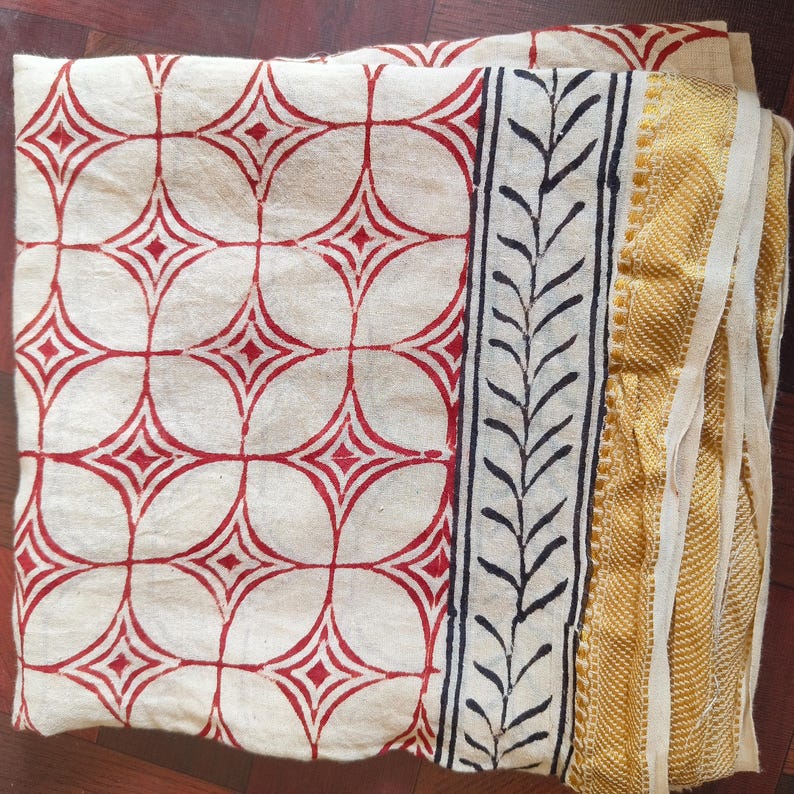 May include: Folded cream-colored fabric with a repeating pattern of red diamonds within circles. A black and white leaf design borders the fabric, with a gold and cream-colored woven edge. The fabric is likely a textile or decorative cloth.