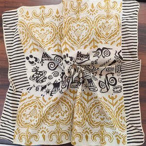 May include: A white cotton square scarf with a black and gold print. The print features a repeating pattern of hearts, swirls, and stylised figures. The scarf has a black and white striped border.