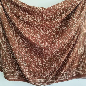 Può includere: Sciarpa marrone con un motivo floreale bianco ripetuto. La sciarpa ha un bordo con un disegno a pois. Il tessuto è drappeggiato, mostrando la consistenza e il movimento del materiale.