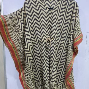 May include: A beige and black chevron patterned kaftan. The kaftan has a V-neck and decorative trim with red, yellow, and green stripes. The sleeves and edges feature an ornate border design. The kaftan is hanging on a black hanger.