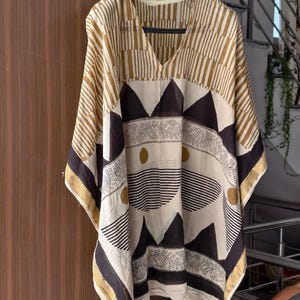 May include: A beige and brown patterned caftan with a V-neck design. The caftan features geometric and floral patterns in shades of brown, black, and gold. The garment is hanging on a black hanger against a wooden door.