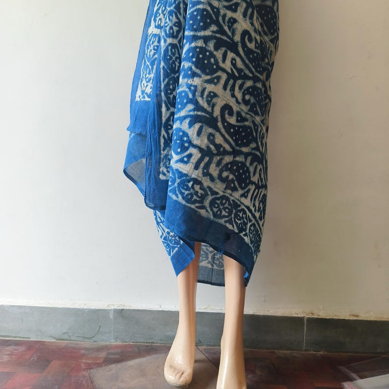 May include: A blue and white patterned scarf with a paisley design. The scarf is draped over a pair of mannequin legs. The fabric has a dark blue border and a repeating pattern of white and blue floral and paisley shapes.