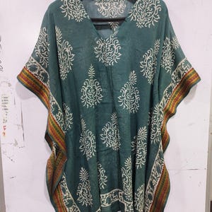 May include: A teal-colored kaftan with a V-neck and short sleeves, adorned with a white floral pattern. The edges feature a decorative border with red, yellow, and green stripes. The garment is hanging on a black hanger.