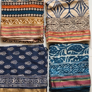 May include: Four folded fabric pieces with intricate block print designs. The fabrics feature a variety of patterns in colors like orange, blue, and beige. Each piece has a distinct border with gold, red, and green accents. The textiles are likely for clothing or home decor.