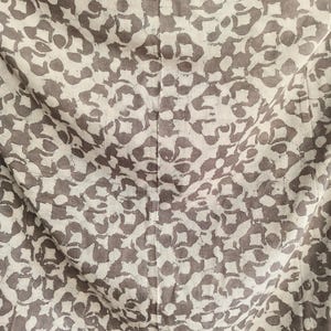 Puede incluir: Primer plano de una tela con un patr&oacute;n repetitivo. El dise&ntilde;o presenta un fondo beige claro con un patr&oacute;n de formas florales de color topo. La tela est&aacute; ligeramente arrugada, con una costura vertical visible en el centro.