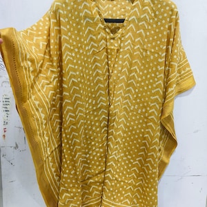 May include: Mustard yellow kaftan with a V-neck and short sleeves. The fabric features a white geometric pattern of dots and chevrons. The edges are trimmed with a gold border. The kaftan is hanging on a black hanger.