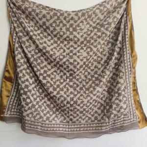 Puede incluir: Bufanda drapeada con un estampado floral repetido en tonos marr&oacute;n y beige. La bufanda tiene un borde con dise&ntilde;o geom&eacute;trico y un ribete dorado. La tela parece suave y fluida.