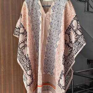 Op de afbeelding: Een perzikkleurige kaftan met een V-hals en een geometrische en paisley print. De kaftan heeft een herhalend cirkelvormig patroon in marineblauw en oranje, met zwart-witte paisley accenten op de mouwen.