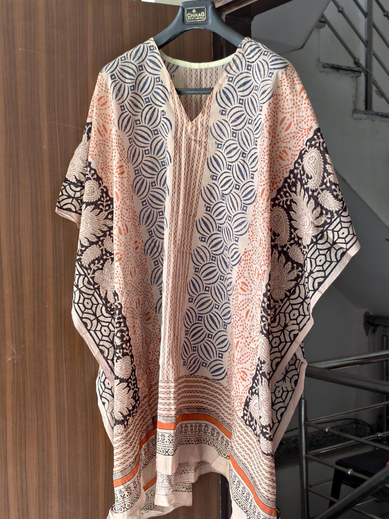 Op de afbeelding: Een perzikkleurige kaftan met een V-hals en een geometrische en paisley print. De kaftan heeft een herhalend cirkelvormig patroon in marineblauw en oranje, met zwart-witte paisley accenten op de mouwen.