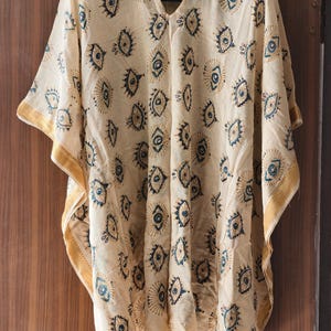 May include: A cream-colored, V-neck kaftan with a repeating pattern of blue and gold eye designs. The edges are trimmed with a gold border. The lightweight fabric hangs loosely.
